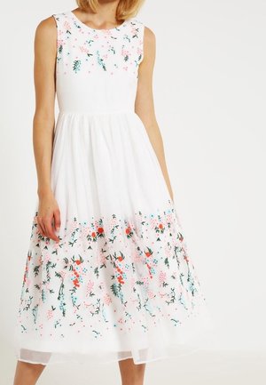 Vestido blanco sin mangas con patrones florales bordados en rosa, naranja y azul en el dobladillo, con una falda fluida y texturizada.