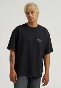 Sort oversized t-shirt lavet af bomuld, med rund hals og korte ærmer; lille logo detalje på venstre bryst. Båret med sorte jeans.