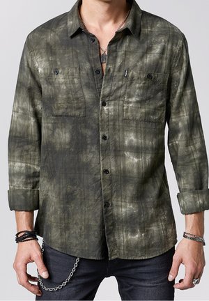 Homme portant une chemise à boutons tie-dye verte et noire avec les manches retroussées, un jean noir, des bracelets et un collier avec un pendentif plaque militaire.