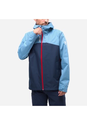 Man zipping up a two-tone blue waterproof jacket with a red zipper and hood, wearing matching navy blue pants.