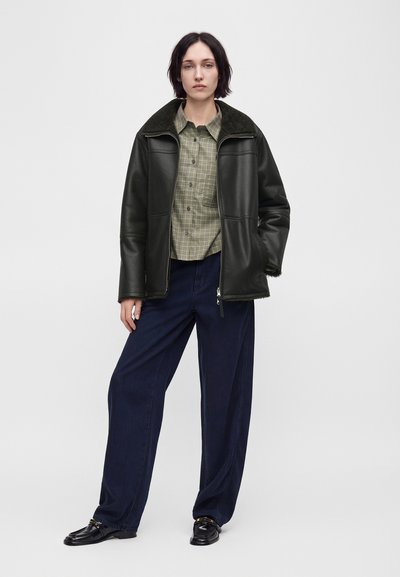 Γυναίκα που φοράει μαύρο σακάκι από shearling, πράσινο καρό πουκάμισο, φαρδιά σκούρα μπλε παντελόνια και μαύρα μοκασίνια, στέκεται μπροστά σε απλό φόντο.