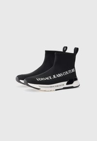 Melni slip-on kedas ar elastīgu augšdaļu, kurām ir balta "VERSACE JEANS COUTURE" zīmola apzīmējums un teksturēta gumijas zole. Dizains ietver vilkšanas cilpas.