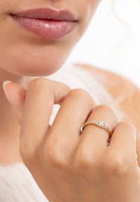 Close-up of lips and hand wearing a gold ring with multiple small diamonds on the ring finger against a light background.