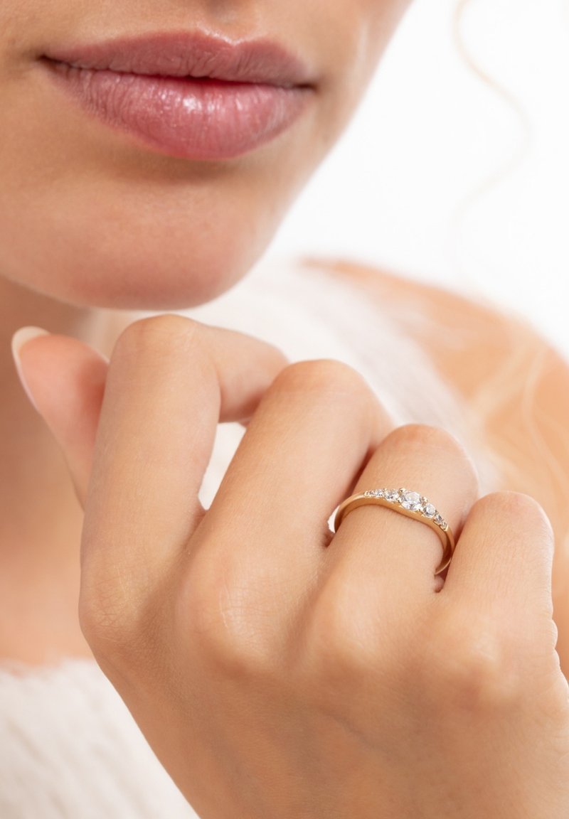 Close-up of lips and hand wearing a gold ring with multiple small diamonds on the ring finger against a light background.