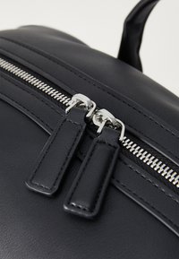 Black faux leather backpack detail featuring a double zip closure with silver hardware and flat zipper pull tabs. Smooth texture, minimalist design.