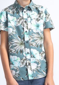 Camicia a maniche corte con bottoni in teal, decorata da grandi motivi floreali bianchi e blu. Presenta una tasca sul petto e bottoni scuri. Tessuto leggero.