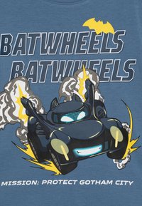 Niebieska koszulka z grafiką rysunkowego pojazdu z akcentami w kształcie nietoperzy, otoczonego dymem i błyskawicami, z napisem poniżej.