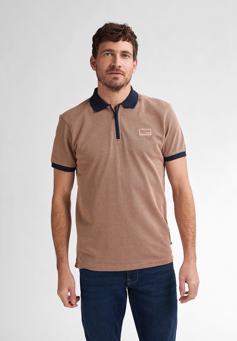 Brun strukturerad pikétröja med dragkedja i kragen, marinblå accenter på kragen och ärmsluten, med ett litet logopatch. Bärs med blå jeans.