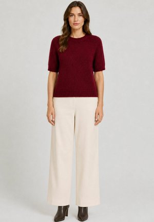 Kvinde iført en kortærmet bordeaux sweater, vidde cremebukser og sorte ankelstøvler, stående mod en ensfarvet lys baggrund.