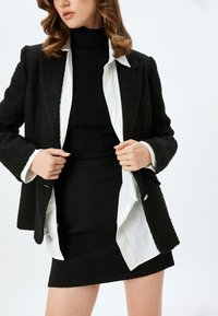 Blazer noir texturé porté sur un turtleneck noir côtelé et une mini jupe noire ajustée, superposé avec une chemise blanche en dessous.