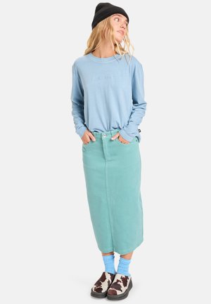 Lichtblauw overhemd met lange mouwen, mintgroene denim rok, blauwe sokken en bruine en witte gevlekte klompen, met een zwarte beanie.
