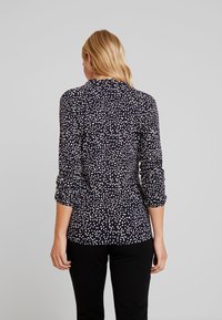 Långärmad marinblå blus med prickigt mönster i vitt och rött. Har rund halsringning och sammangående ärmar, tillverkad av lättviktsmaterial.