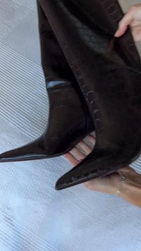 Fekete krokodilbőr mintájú, hegyes orrú, fényes felületű magas sarkú csizma, amelyet egy texturált világos háttér előtt tartanak.