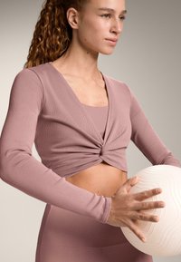 Rebrast, dolg rokav mauve skrajšan zgornji del z detajlom zasuka spredaj, nošen čez ujemajočo se oprijeto majico, v kombinaciji s teksturirano belo kroglico.