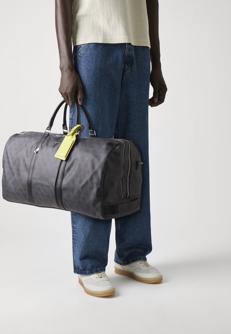 Sac de sport noir avec un motif subtil, poignées robustes, une étiquette jaune et une fermeture éclair, tenu par une personne en jean bleu et chaussures blanches.