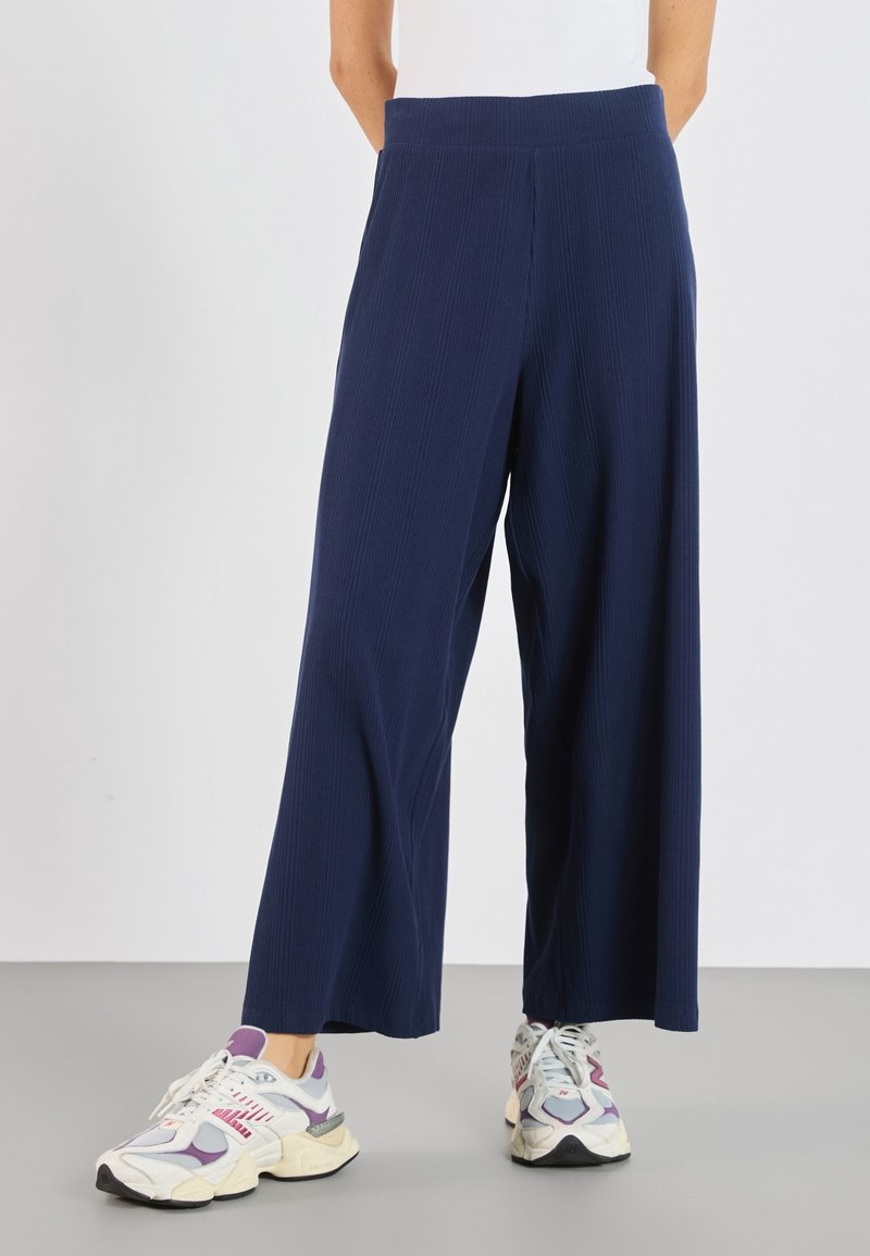 Personne portant un pantalon large côtelé bleu marine et des baskets blanches avec des accents violets, debout sur un sol gris.