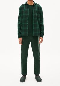 Uomo che indossa una camicia a quadri verde scuro con bottoni sopra una maglietta verde, pantaloni verde scuro con coulisse e scarpe slip-on verdi su sfondo bianco.