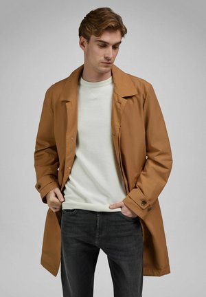 Junger Mann trägt einen braunen Trenchcoat über einem weißen Pullover und schwarzen Jeans, steht vor einem schlichten grauen Hintergrund.
