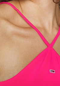 Fuksjowy top na ramiączkach z cienkimi paskami, z małym emblematem logo w czerwono-biało-niebieskich akcentach po lewej stronie. Miękki, elastyczny materiał.