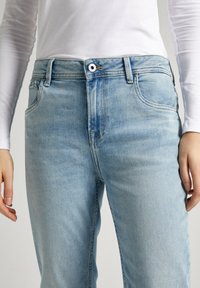 Svetlomodré rifle z denimu so slabým vyblednutím, s piatimi vreckami a všetkým zapínaním na gombík. Materiál je mäkký s hladkou textúrou.