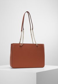 Sac fourre-tout en cuir marron avec deux poignées, l'une d'elles ornée d'une chaîne. Forme rectangulaire avec une texture lisse et un design minimaliste.