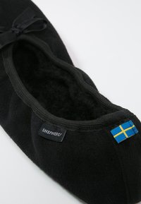 Black felt slipper with rounded shape, ribbed trim, and a bow. Features a label and a small Swedish flag accent on the side.