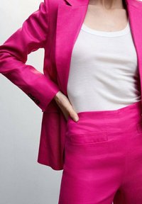 Fuksjowy, dopasowany żakiet i spodnie wykonane z gładkiego materiału, w połączeniu z białym, prążkowanym topem na ramiączkach. Żakiet ma zapinane mankiety, a spodnie mają kieszenie.
