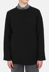 Schwarzer gerippter Pullover mit rundem Halsausschnitt und langen Ärmeln. Weicher Stoff mit lockerer Passform, getragen über einem gestreiften grauen und weißen Hemd.