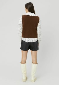 Femme se tenant de dos, portant un pull sans manches marron, une blouse à fleurs, un short noir et des bottes blanches montantes contre un fond uni.