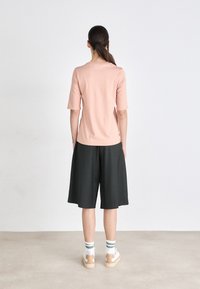 Svetloružový top s krátkym rukávom skombinovaný s tmavoškórovými culottes, bielymi a zelenými pruhovanými ponožkami a béžovými teniskami. Jednoduché pozadie.