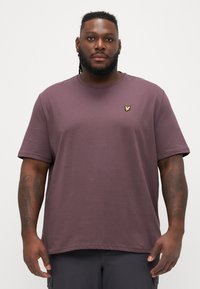 Lila Kurzarm-T-Shirt mit Rundhalsausschnitt. Es hat ein kleines gelbes Logo auf der linken Brust. Der Stoff wirkt weich mit einer glatten Textur.