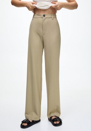 Person trägt beige hoch taillierte, weit geschnittene Hose und schwarze Sandalen zum Hineinschlüpfen, hebt ein weißes, strukturiertes Oberteil an, um den Bauch freizulegen, vor einfachem Hintergrund.