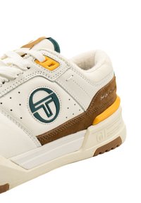 Baltas sneakers ar brūnām zamšādas detaļām, tīlzaļa logo un dzeltenu aci. Funkcijas ietver perforētas detaļas un teksturētu zoli labākai saķerei.