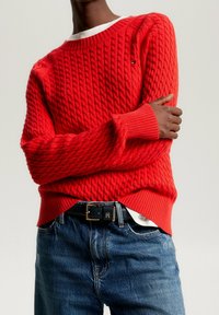 Personne portant un pull en maille rouge vif par-dessus une chemise blanche, associé à un jean bleu et une ceinture noire avec une boucle dorée.