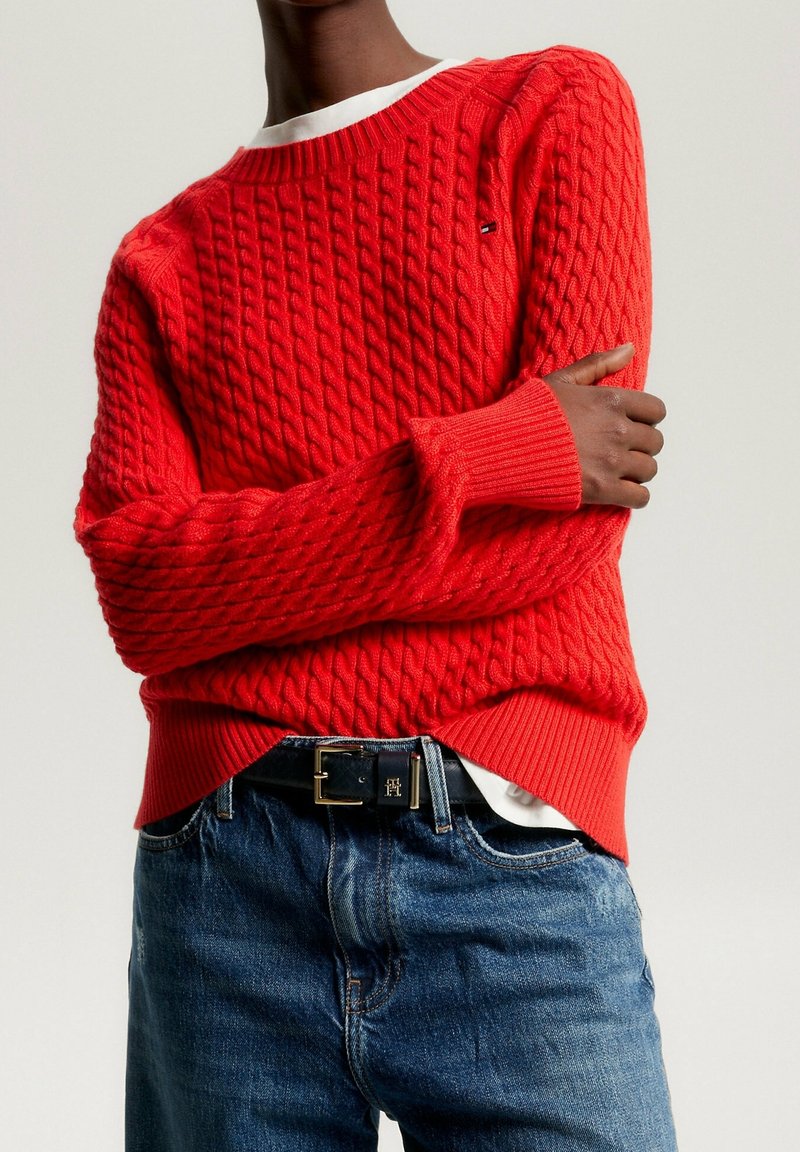 Personne portant un pull en maille rouge vif par-dessus une chemise blanche, associé à un jean bleu et une ceinture noire avec une boucle dorée.