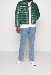 Chaqueta acolchada verde con acabado brillante, cierre con cremallera y cuello alto, combinada con una sudadera blanca, vaqueros azul claro y zapatillas blancas.