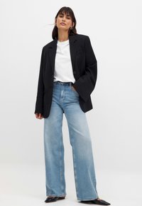 Čierny oversize blazer, biele tričko a široké svetlomodré džínsy. Čierne špicaté topánky. Hladká tkanina so štruktúrovanými ramenami a voľným strihom.