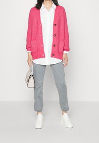 Rosa stickad cardigan med ribbad textur, framfickor och bruna knappar, lager på en vit skjorta, ihop med grå byxor och vita sneakers.