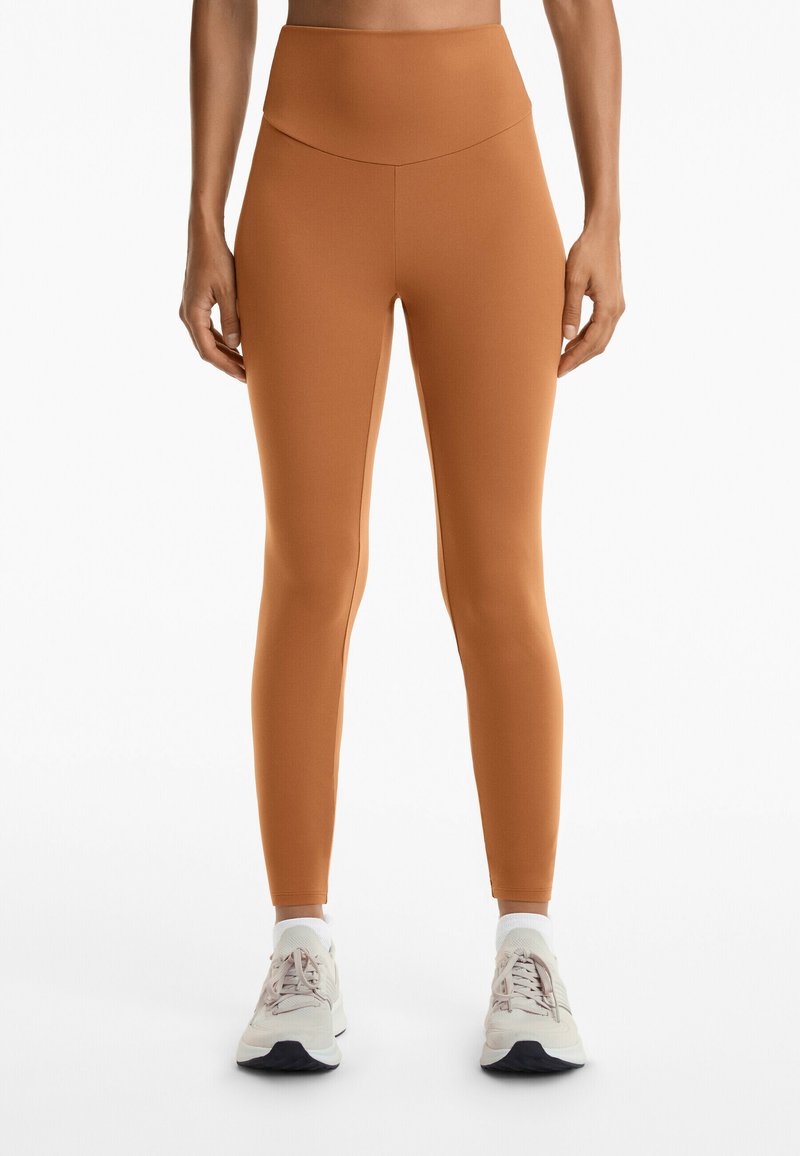 Högt skurna, figurnära leggings gjorda av stretchigt tyg i en varm brun nyans, kombinerade med ljusa sneakers med grå och vita detaljer.