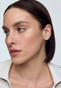 Złote kolczyki wkręty z małym wzorem niedźwiadka. Modelka ma proste, ciemne włosy i nosi jasnobeżową kurtkę z czarnym wykończeniem.