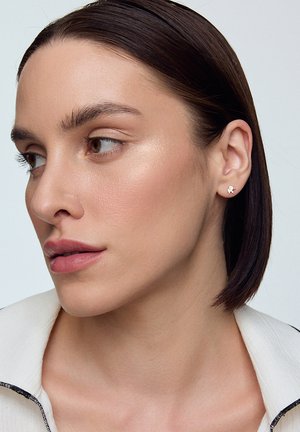 Złote kolczyki wkręty z małym wzorem niedźwiadka. Modelka ma proste, ciemne włosy i nosi jasnobeżową kurtkę z czarnym wykończeniem.