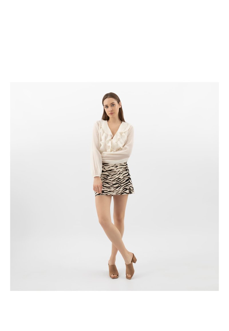 Donna in piedi con gambe incrociate, che indossa una camicetta bianca con balze, una minigonna a stampa zebra e sandali marroni con tacco su uno sfondo bianco semplice.