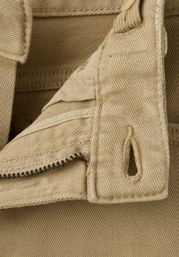 Prim-plan cu o țesătură bej, cu fermoar, nasture și buzunar cusut având o suprafață texturată. Include un detaliu mic cu o fantă.