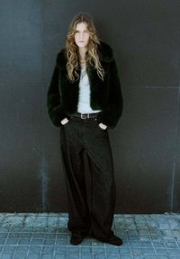 Veste courte en fausse fourrure noire, haut blanc moulant et pantalon en jean noir à jambes larges. Le mannequin se tient sur un sol carrelé, expression neutre, texture naturelle.