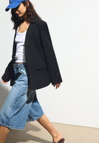 Čierny blejzer s voľným strihom, skombinovaný s bielym topom bez rukávov a širokými modrými džínsovými culottes. Osoba má na sebe hnedé sandále a modrú čiapku.