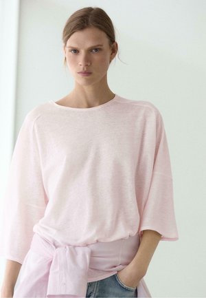 Kvinde med lyst brunt hår iført en løs, lyserød top og matchende skjorte bundet ved taljen, står med den ene hånd i lommen på blå jeans.