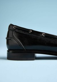 Schwarzer Leder-Slipper mit elegantem Design, versehen mit Metallösen, niedrigem Blockabsatz und glatter Oberfläche. Seitenansicht vor blauem Hintergrund.