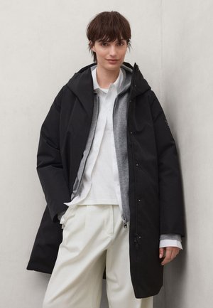 Persona con cabello corto y castaño que lleva un abrigo negro con capucha, un suéter gris con cremallera, una camisa blanca y pantalones de color crema, de pie junto a una pared clara.