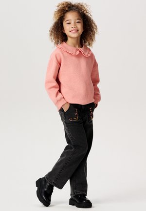 Hellrosa Sweatshirt mit Rüschensaum, kombiniert mit schwarzen Jeans mit Blumenstickerei. Abgerundet mit schwarzen klobigen Stiefeln.
