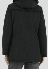 Person som bär en svart huva jacka med knappdetalj på ryggen, kombinerad med rutiga byxor, vänd bort från kameran mot en enfärgad bakgrund.